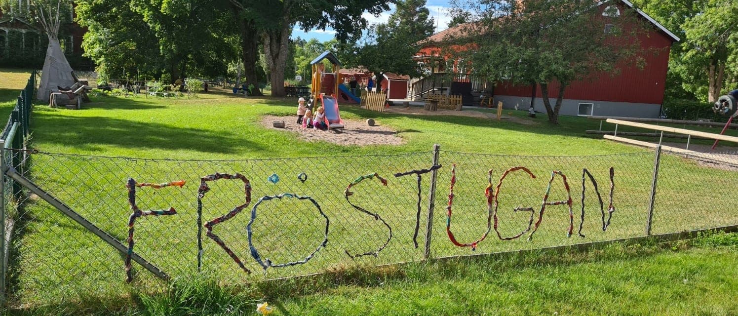 Förskolans gård en sommardag. Namnet FRÖSTUGAN är flätat i tygremsor genom stängslet framför gräsmattan, lekställningen och den röda skolbyggnaden.